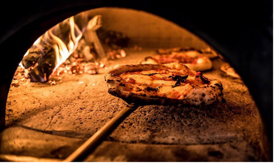 A color image showing a wood-fired pizza oven with a pizza being baked inside, flames visible in the background at Mozzeria.