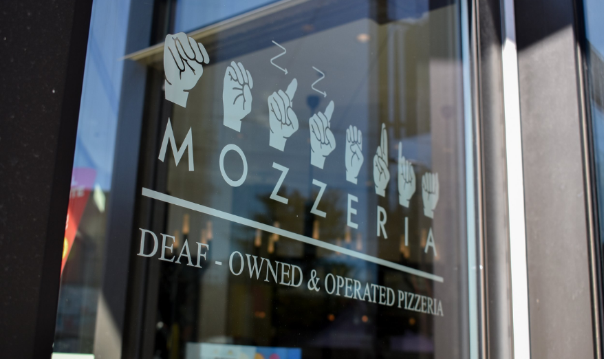 A color close-up of the Mozzeria sign on the window, indicating "Deaf-Owned & Operated Pizzeria" with the reflection of the street visible.