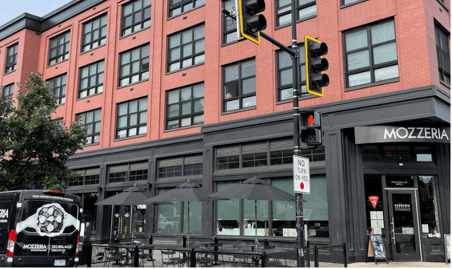 A color image of the exterior of Mozzeria, a restaurant in a red brick building with large windows and black trim. The Mozzeria sign is visible above the entrance, and a Mozzeria delivery van is parked nearby. There are also black umbrellas set up outside for outdoor dining, and a traffic light and "No Turn on Red" sign are in the foreground.